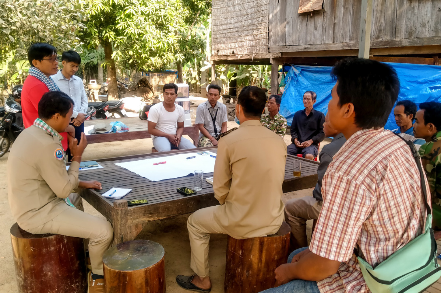 A group of people is sitting around a wooden table on tree stump stools, brainstorming and recording ideas and objectives on a poster in the centre of the table.