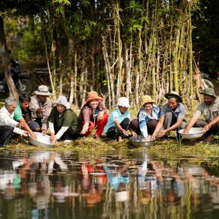 A group of nine community members waring hats is sitting in a squatting position in front of a pond. They are releasing small fingerlings from a bowl into the pond.