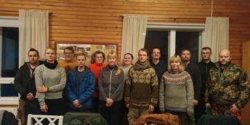 A group of 13 people looking into the camera . They appear to be inside, the walls are made out of wood. They are all wearing warm clothes, it must be cold outside.