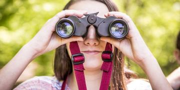 A young girl peers through binoculars that are pointed at the viewer. 