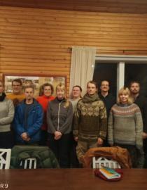 A group of 13 people looking into the camera . They appear to be inside, the walls are made out of wood. They are all wearing warm clothes, it must be cold outside.