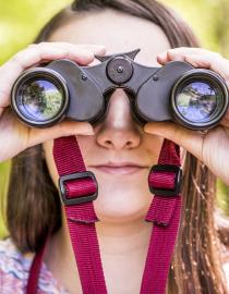 A young girl peers through binoculars that are pointed at the viewer.